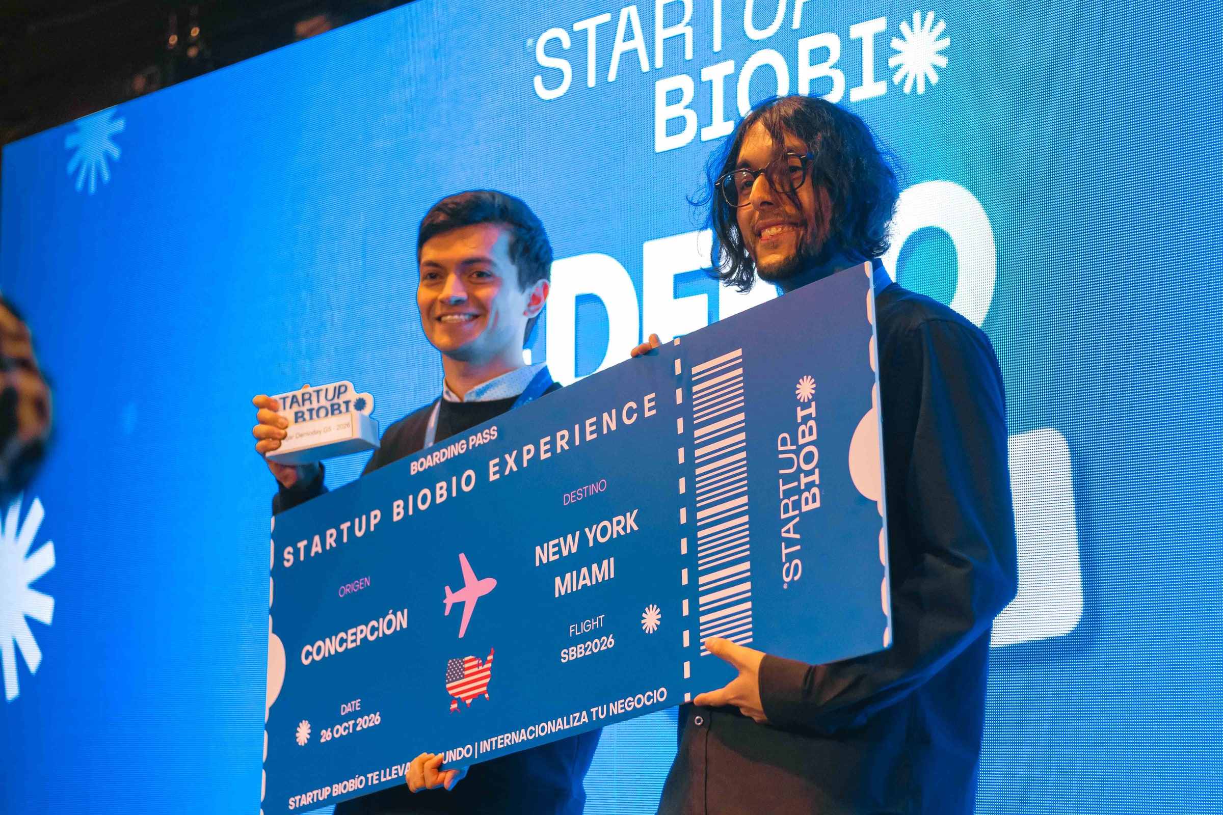 Two PAIR team members on the stage of the Startup Biobío final holding the symbolic prize ticket, a flight from Concepción to New York and Miami.